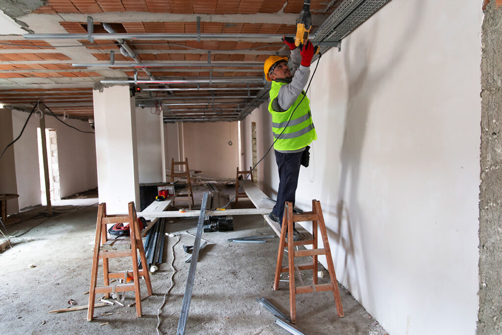 construction worker on a platform indoors working overhead installing drywall and using a drill
